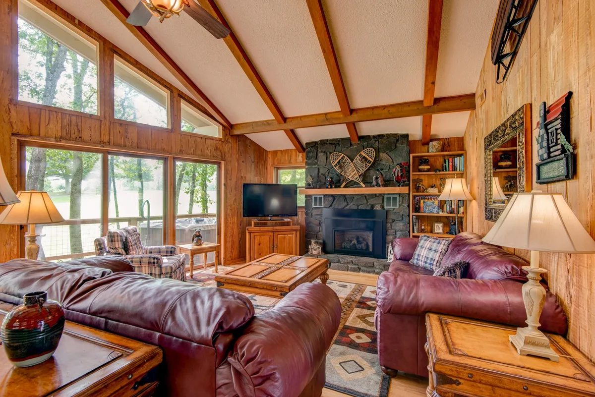 A comfortable and spacious living room with oversized leather sofas arranged around a striking stone fireplace. Large windows flood the area with natural light, while wooden furniture and decor invite guests to relax, socialize, or enjoy a movie night together.