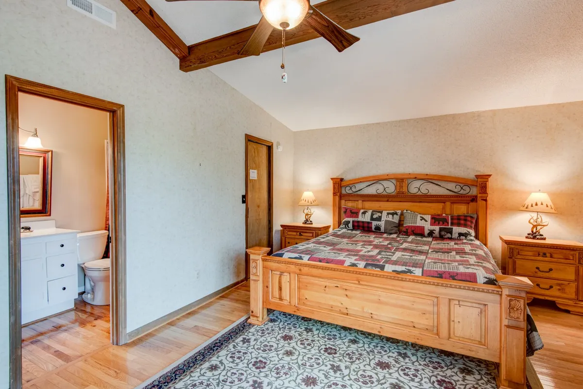 A warmly lit bedroom featuring a wooden bed frame with a decorative quilt, complemented by matching nightstands with lamps and a visible bathroom door. The soft color palette and mountain-inspired accents create a cozy, restful atmosphere ideal for relaxation and privacy.