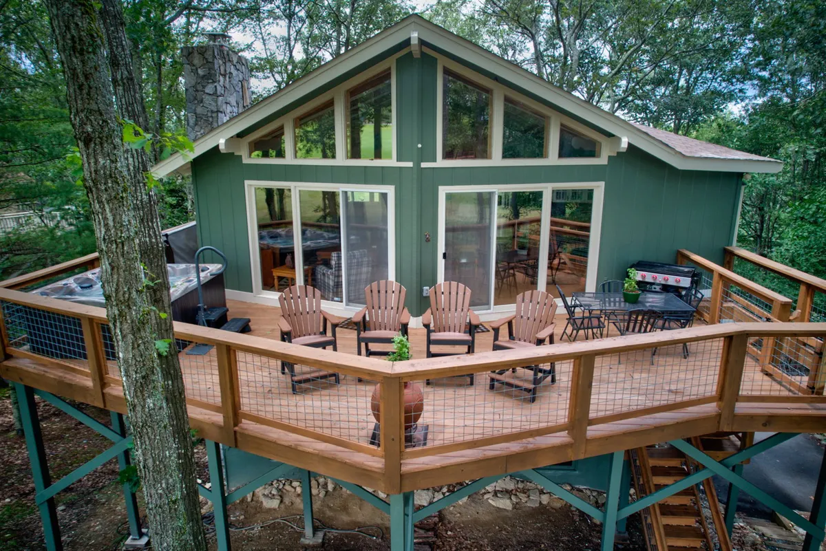 The image showcases the welcoming exterior of a mountain home with a spacious deck featuring Adirondack chairs, large windows that hint at bright, open interiors, a striking stone chimney, and an outdoor grill. The setting suggests relaxing with loved ones, taking in sunset views, and enjoying the serene beauty of the surrounding golf course and mountains.