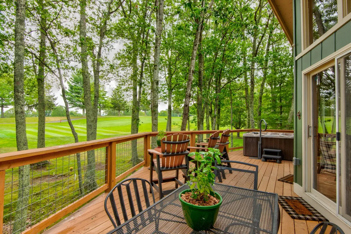 This photo showcases another angle of a wooden deck featuring an outdoor dining set and the hot tub, set against a lush green backdrop. The scene conveys relaxation and entertainment, with space for both dining and soaking. The view from the deck offers further connection to the outdoors and the golf course, highlighting options for both sunrise breakfasts and evening stargazing.