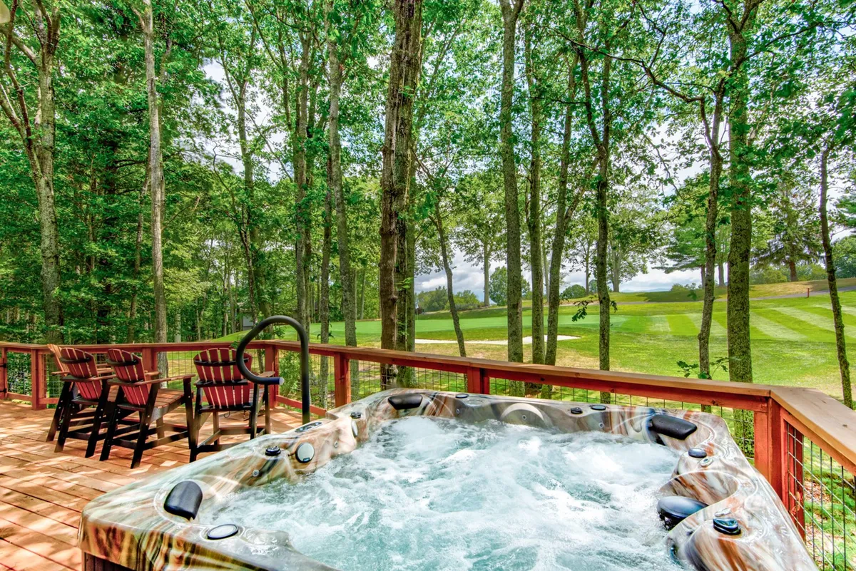 This exterior image highlights a spacious wooden deck with a hot tub in the foreground, surrounded by lush trees and a clear view of the nearby golf course. The deck is equipped with comfortable seating, perfect for relaxing with a drink and taking in tranquil golf course vistas. The atmosphere feels peaceful, inviting guests to enjoy stargazing, morning coffee, or evening barbecues overlooking the green.