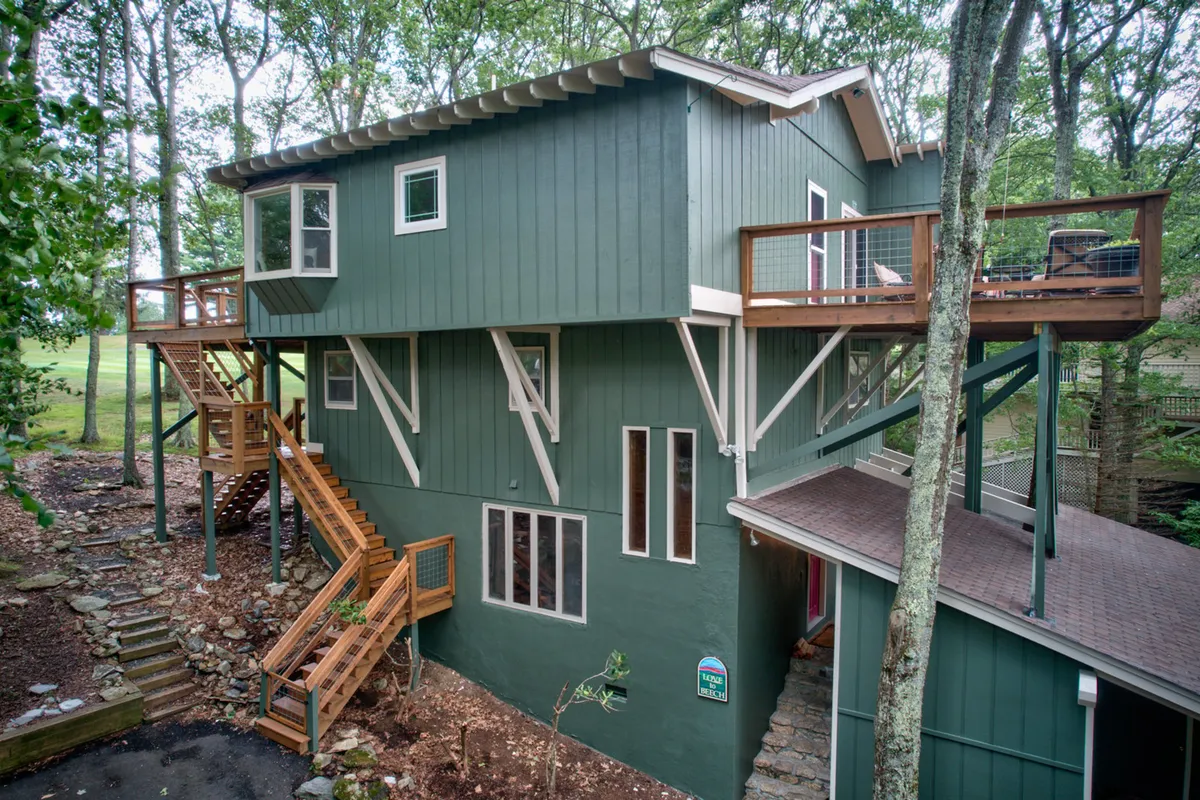 This image shows the multi-level structure of the home surrounded by lush trees, with visible wooden staircases and expansive balcony areas. The angle emphasizes the west-facing balcony where sunset views can be enjoyed, and the welcoming outdoor spaces that invite relaxation or socializing.