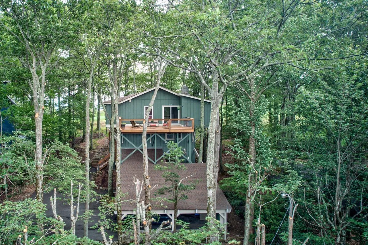 A tranquil, treehouse-style home with a wooden deck nestled among tall trees and a driveway leading up, blending seamlessly into the forested environment. The setting celebrates the home’s close relationship with the surrounding mountains and outdoor recreation.