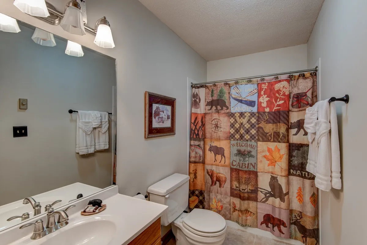 A rustic, inviting bathroom showcasing a wildlife- and cabin-themed shower curtain, a bathroom vanity with sink, towels on the rack, and framed artwork. The space is well-lit and thoughtfully decorated, providing a sense of comfort and post-adventure relaxation.