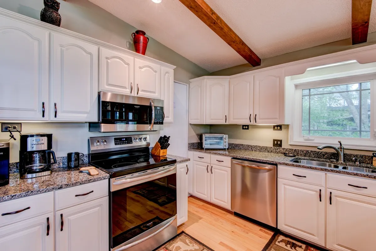A warm, inviting kitchen with stainless steel appliances gleaming beside a granite-topped island, wooden beams overhead, and a sunlit kitchen window. The space is arranged for cooking, entertaining, and gathering, with visible elements suggesting room for multiple guests to comfortably congregate.
