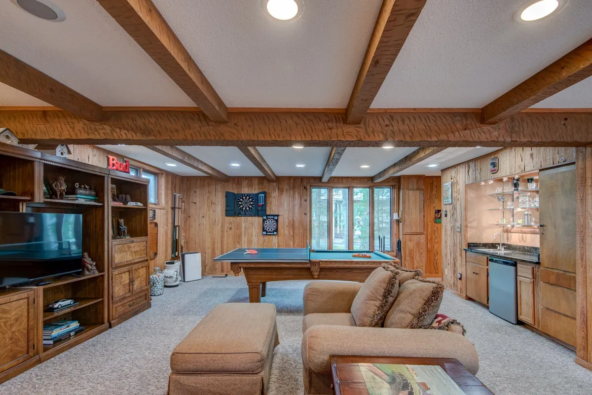A stylish and spacious game room featuring a central pool table, cozy seating area with sofas, an entertainment center with TV, and a bar area in the background. The room is well-lit and designed for social gatherings and friendly competition.