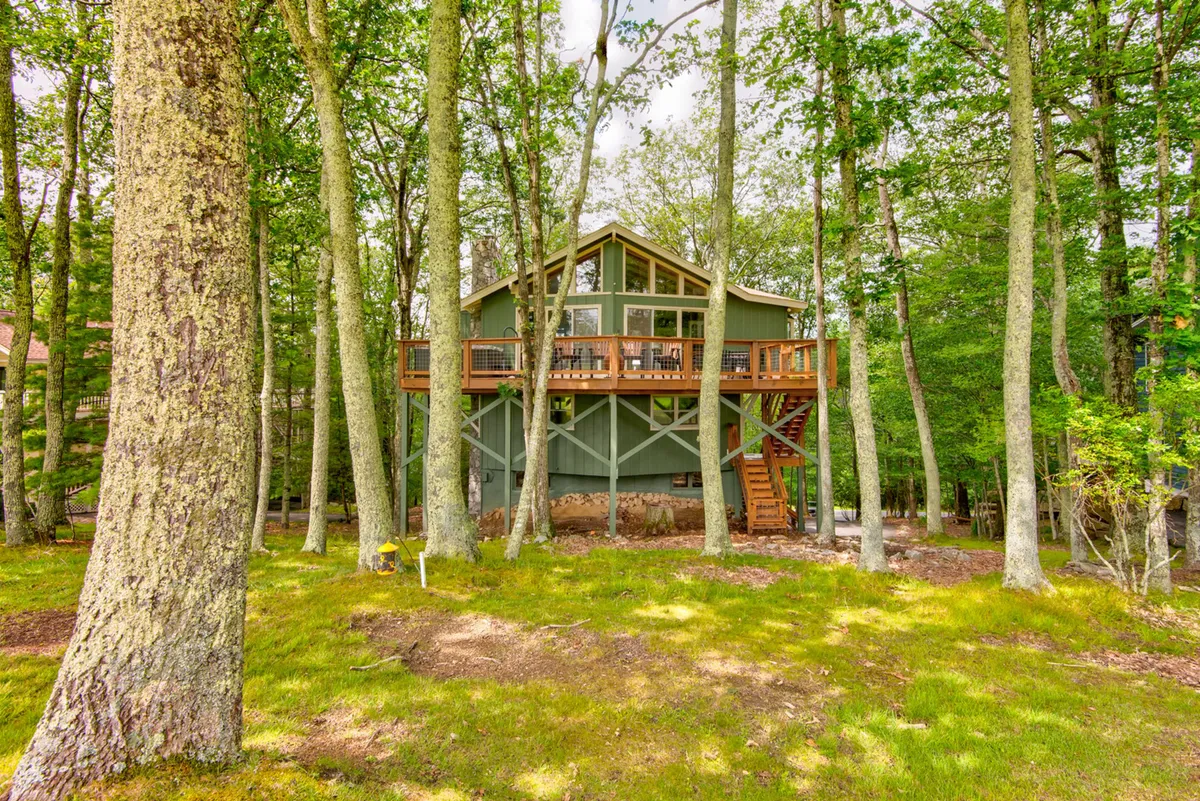 An elevated house with a spacious wooden deck nestled among green trees, featuring comfortable seating that overlooks the landscape. The view invites outdoor dining or simply relaxing while surrounded by nature—perfect for watching sunsets or enjoying tranquil evenings.