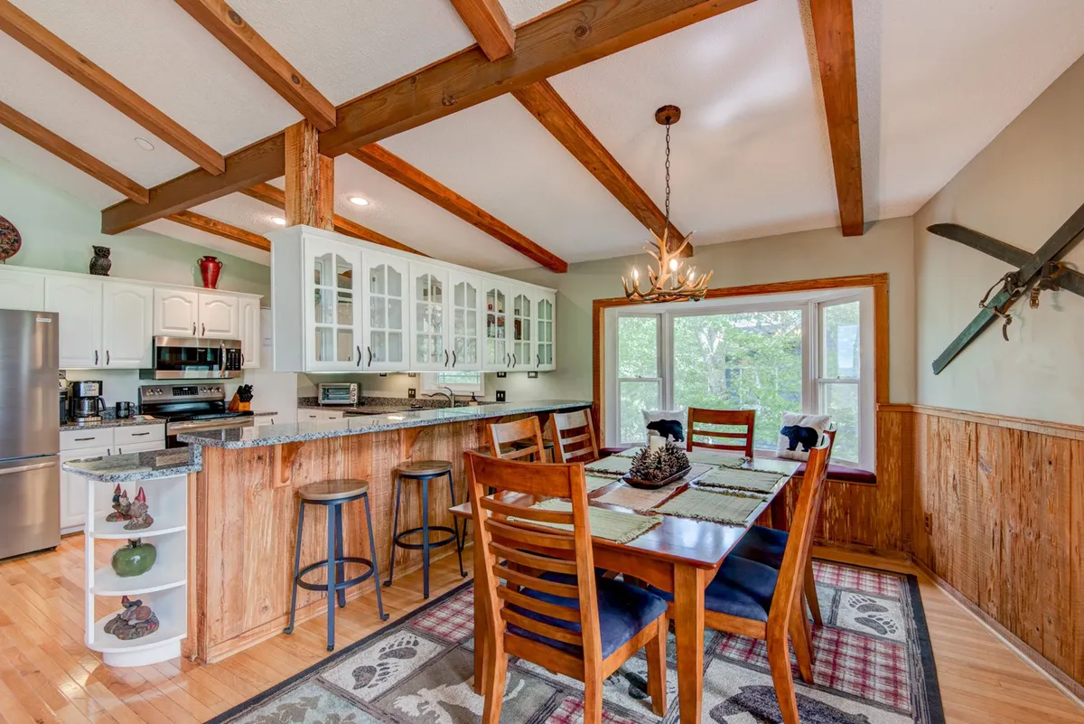 A spacious dining area seamlessly connected to the kitchen, featuring a large dining table with seating for six, a kitchen island with additional barstools, and generous natural light pouring in through a large window. The wooden beams add mountain charm, and the setup emphasizes communal dining and casual gatherings.