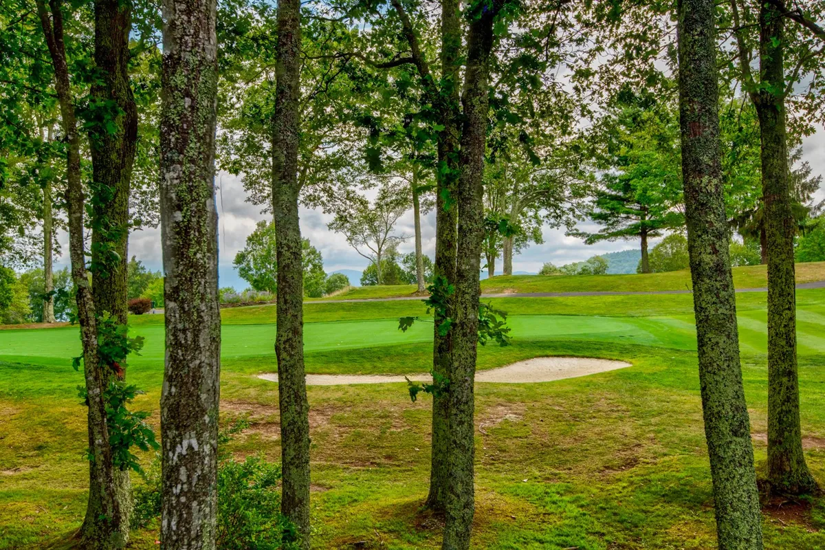 A picturesque view of the Beech Mountain Golf Course green with a sand trap in the foreground, surrounded by mature trees and lush grass. The perspective places the viewer directly on the course, capturing the relaxed, scenic environment perfect for golfers.