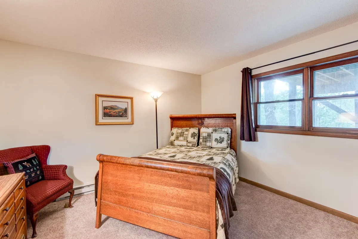 A cozy queen bedroom with a wooden bed, plaid bedding, framed artwork, and a window that lets in natural light. The warm tones and soft furnishings enhance the feeling of a restful private oasis.