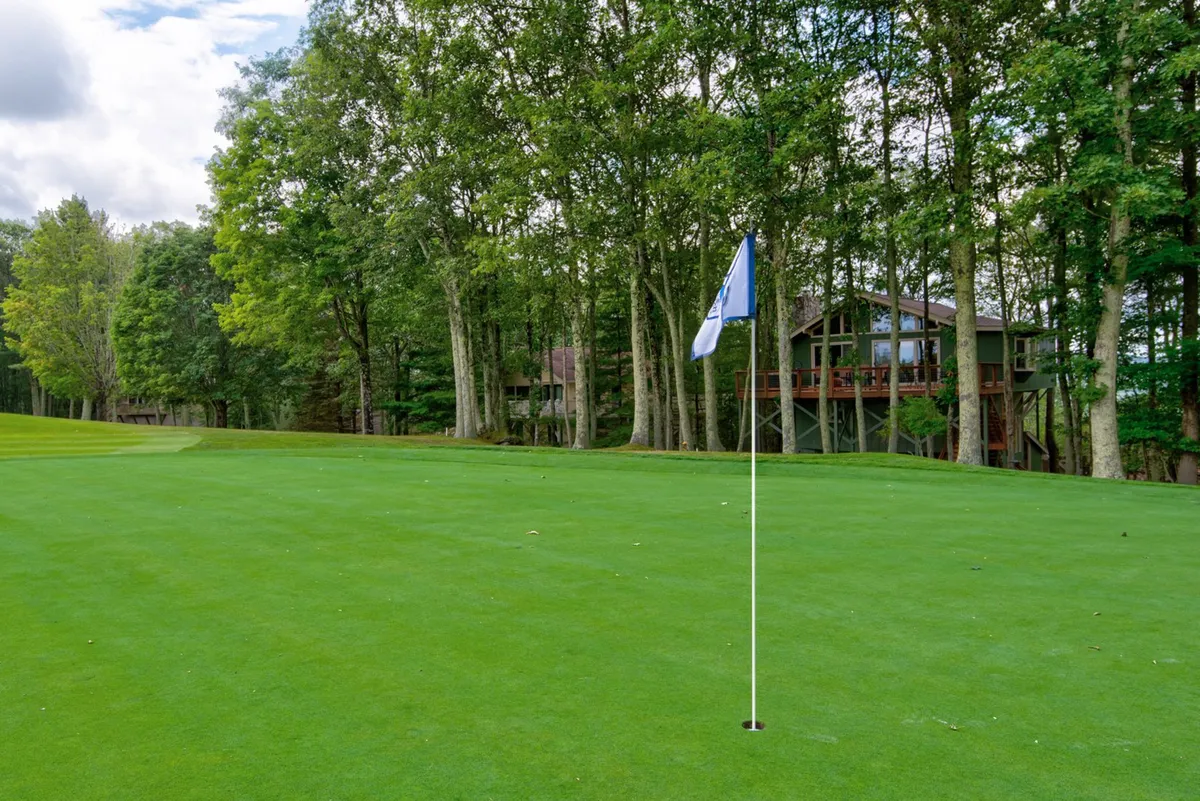 A scenic shot of a manicured golf course flag surrounded by lush grass and trees, with a house in the background. The space is open and inviting, perfect for group outings and enjoying mountain air.