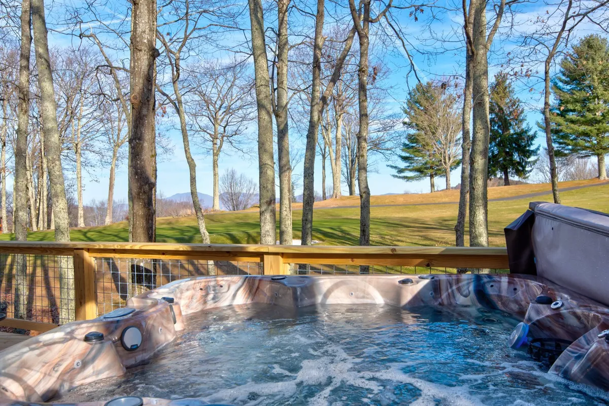 A view of the exterior deck with a bubbling hot tub overlooking a serene mountain landscape, surrounded by trees. The scene is set at sunset, with warm light casting a relaxing glow over the area, inviting groups to unwind while socializing or enjoying the natural setting.